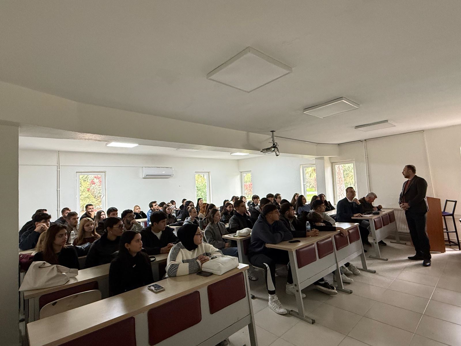 A group photo taken in the garden of Dalaman Vocational School. Faculty members and students stand together in front of an Atatürk bust, posing for the camera. The school building and tall pine trees are visible in the background. The large group is smiling while standing outdoors.